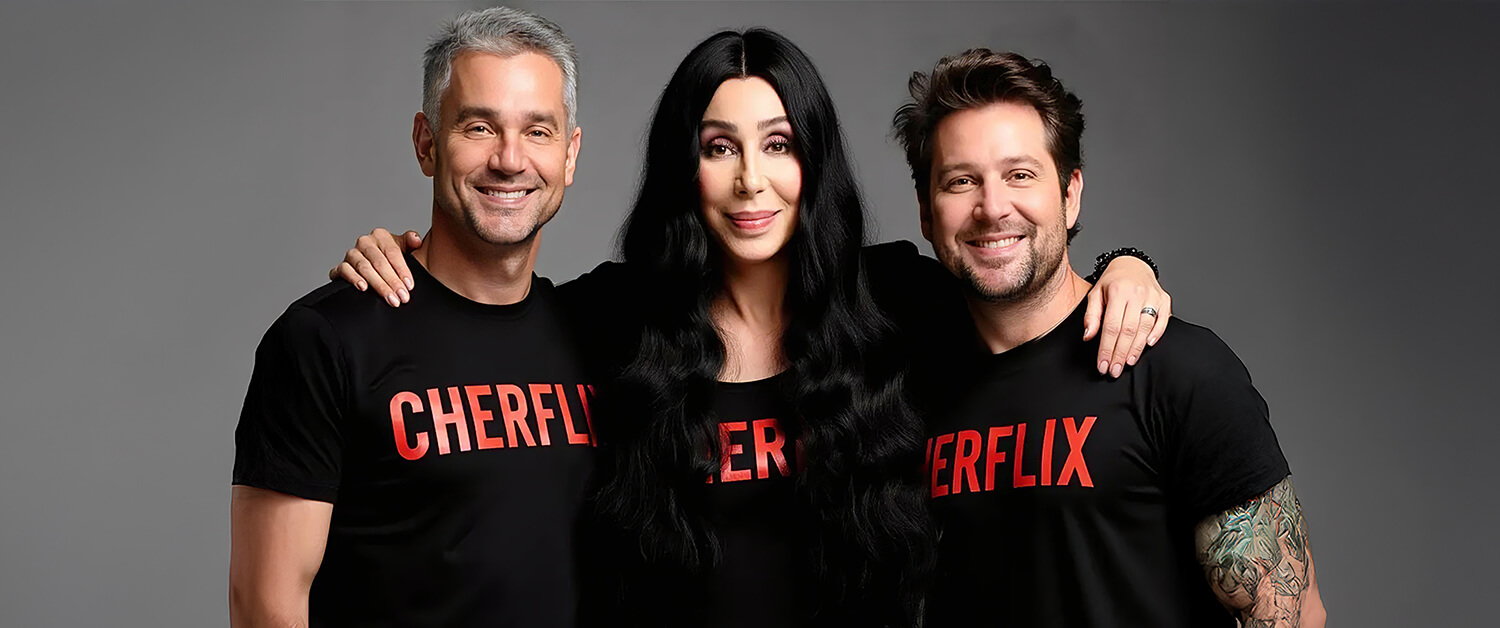 Cher stands between two smiling male fans, all wearing black T-shirts with "CHERFLIX" in red lettering, against a gray background. She has long wavy black hair, and her arms are draped around their shoulders.